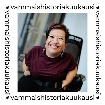 Anna ler brett och sitter i en elektrisk rullstol. Anna har kort hår med färgade ränder, smink på ögonen och stora örhängen. Bilden är omgiven av en vit ram där det står ”vammaishistoriakuukausi”.