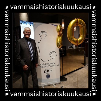 Håkan står bredvid en roll-up från Finlandssvenska teckenspråkiga rf, iklädd kostym och slips. Det finns också guldfärgade folieballonger med nummer 2 och 0 i närheten. Håkan har vitt hår och glasögon. Bilden är omgiven av en svart ram där det står ”vammaishistoriakuukausi”.