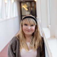 Sanni Virta on pohjoismaisen näköinen, vaaleapiirtenen ja -hiuksinen nainen, joka hymyilee Kaapelitehtaan Huokausten sillalla.