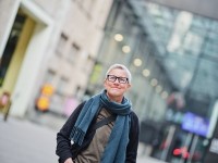 Rita is outside, looking up and past the camera. She has very short light gray hair, glasses, a shoulder bag. A warm scarf, a black cardigan. In the background, the Cable Factory wall and the Glass Courtyard are blurred.