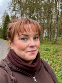Eeva photographed outdoors from the shoulders up. Brown hair tied up, bangs. Scarf around her neck.