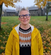 Rita står utomhus en höstdag, iklädd tröja och gul regnrock. I bakgrunden syns lönnar och gamla stenhus.