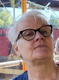 Marjo has short white hair and black-rimmed glasses. The photo was taken on a sunny summer day.